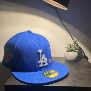 LA Dodgers 59FIFTY keps - Säljer en blå LA Dodgers 59FIFTY keps från New Era med broderad LA-logga framtill, 1988 World Series patch på sidan och texten 'Love & Hustle = Game'. Platt skärm i grått och klassisk basebollstil. Perfekt för dig som vill sticka ut med sportig vibe.