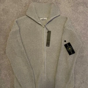 Grå stickad kofta från Stone Island - Säljer en grå stickad kofta från Stone Island med dragkedja och hög krage. Klassisk badge på vänster ärm och grovstickad struktur som ger en schysst vibe. Perfekt för dig som gillar streetwear och vill ha något varmt och stilrent.