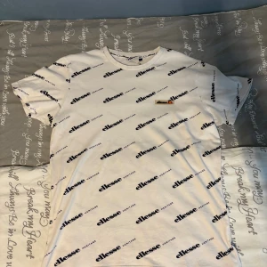 Vit t-shirt med Ellesse-logga, M - Vit t-shirt från Ellesse i storlek M med svart logotyptryck över hela plagget. Klassisk rund halsringning och korta ärmar. Tillverkad i mjuk bomull för en skön känsla. Perfekt för dig som gillar streetwear och sportig stil.
