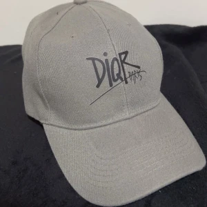 Grå keps från Dior Paris - Snygg grå keps från Dior Paris med svart graffiti-inspirerad logga framtill. Klassisk böjd skärm och justerbar rem baktill för perfekt passform. Tillverkad i slitstark bomull som ger en cool och urban vibe.