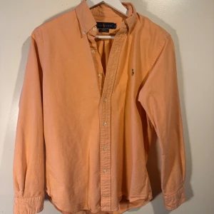 Orange slim fit skjorta från Ralph Lauren - Fräsch orange skjorta från Ralph Lauren i slim fit stretch oxford. Klassisk button-down krage, broderad logga på bröstet och långa ärmar med knappar vid manschetten. Skjortan har en mjuk bomullskänsla och passar perfekt till jeans eller chinos.