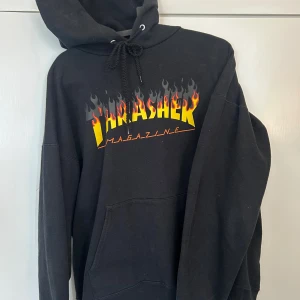 Svart Thrasher hoodie med flames - Svart hoodie från Thrasher med ikoniskt flames-tryck i gult och rött på bröstet. Klassisk känguruficka, snörning i huvan och mjukt material. Perfekt för dig som gillar streetwear och skatekultur.