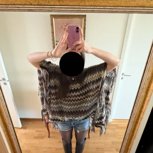 Missoni inspirerad poncho  med fransar - Säljer en poncho-inspirerad topp med zigzag-mönster i brunt, beige, grått och svart. Toppens loose fit och fransar längs kanterna ger en chill och bohemisk vibe. Perfekt att slänga över en hoodie eller t-shirt för extra lager. Materialet känns stickat och mjukt.