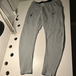 Grå Nike LFC mjukisbyxor - Snygga grå mjukisbyxor från Nike med Liverpool FC-logga på ena benet och svart Nike Swoosh på andra. Byxorna har elastisk midja, dragkedjeficka och är tillverkade i mjukt bomullsmaterial. Perfekta för chill eller träning.
