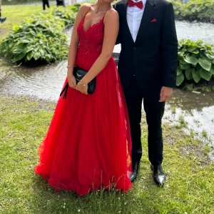 Röd långklänning med spets och öppen rygg - Säljer en röd långklänning med tunna axelband, spetsdetaljer upptill och v-ringning. Klänningen har en öppen rygg och ett flygigt, voluminöst kjolparti i tyll. Perfekt för bal eller student! Snygg och elegant look.