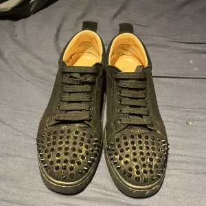 Svarta sneakers med nitar från Louboutin - Säljer ett par svarta sneakers från Christian Louboutin med klassisk röd sula. Skorna har svarta nitar över tån, snörning och är tillverkade i mocka. Riktigt edgy och sticker ut med sin coola design.