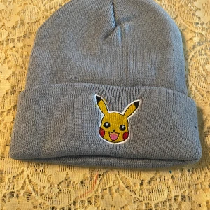 Grå Pikachu mössa stickad - Säljer en grå stickad mössa med broderad Pikachu på uppviket. Perfekt för dig som älskar Pokémon och vill ha en mysig accessoar till vintern. Mössan är mjuk och har en klassisk form med uppvikt kant.  Barn Stlk 
