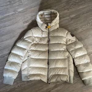 Vit pufferjacka från Parajumpers - Snygg vit pufferjacka från Parajumpers med hög krage, huva och coolt patch på ärmen. Jackan har dragkedja framtill, två sidofickor med dragkedja och ribbade muddar. Materialet är glansigt och känns riktigt lyxigt. Perfekt för kalla dagar. 100% äkta, hör av er vid minsta fundering.