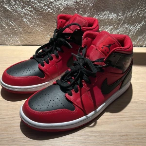Nike Air Jordan 1 Mid Röd/Svart 42 - Nike Air Jordan 1 Mid i rött och svart med klassisk svart swoosh och Air Jordan-logga på sidan. Skorna har svarta snören, vit mellansula och röd yttersula. Tillverkade i skinn och syntet, med perforerad tå och högt skaft för ikonisk look.