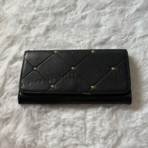 Svart quiltad plånbok med nitar - Snygg svart plånbok med quiltad framsida och små guldfärgade nitar. Plånboken har flera fack för kort, sedlar och mynt, samt en dragkedja med gulddetalj. Tillverkad i skinnliknande material med stilren look.