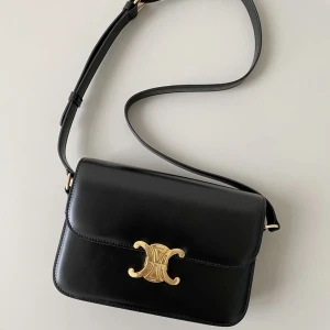 Svart Celine Triomphe axelväska - Snygg svart axelväska från Celine, modell Triomphe, med stilren rektangulär form och justerbar rem. Väskan är gjord i slätt skinn och har ett ikoniskt guldspänne framtill med Celine-logga. Perfekt för dig som gillar klassisk och lyxig design.