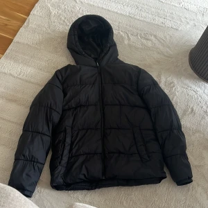 Svart pufferjacka från Jack & Jones - Svart pufferjacka från Jack & Jones i storlek L. Jackan har huva, dragkedja framtill och två sidofickor. Den är quiltad och har en loose passform, perfekt för kalla dagar. Materialet är syntet och jackan är långärmad.