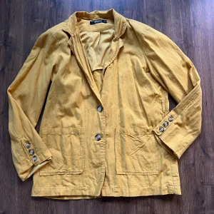 Senapsgul kavaj  - Senapsgul kavaj i 90% “more sustainable” bomull och 10% linne. Har två fickor framtill samt fina knappar. Kavajen har lätt / diskret axelvaddering.