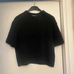 Svart stickad tröja från Gina Tricot - Svart stickad tröja från Gina Tricot i croppad modell med korta ärmar och rund hals. Mjuk och fluffig känsla, perfekt för lager-på-lager under höst och vinter. Stilren och enkel design som funkar till många olika looks.