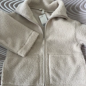 Beige teddyjacka från H&M, strl XS - Supermjuk och mysig teddyjacka i beige från H&M. Jackan har hög krage, två stora fickor framtill och stängs med dragkedja. Perfekt för kyliga dagar och ger en chill vibe till din outfit.