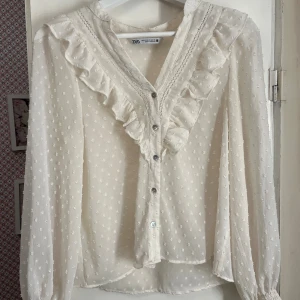 Vit blus med volanger från Zara - Supersöt vit blus från Zara med volanger längs v-ringningen och knappar framtill. Blusen har ett diskret prickigt mönster och spetsdetaljer. Kommer inte till användning längre!🙌🏼