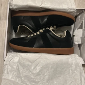 Maison Margiela gats svarta  - Snygga svarta sneakers med klassisk låg profil och kontrasterande brun gummisula. Skorna har vita snören och är tillverkade i skinn med en rund tå. Perfekta för dig som gillar stilrena och tidlösa sneakers. ¡OBS! Skorna är oanvända men boxen som kommer med är sliten!
