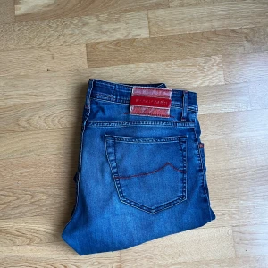 Blå jeans från Jacob Cohen - Snygga blå jeans från Jacob Cohen med röda sömmar och detaljer. Klassisk femficksmodell med raka ben och läderpatch bak i midjan. Jeansen är tillverkade i mjukt denimtyg som ger en skön känsla och stilren look.
