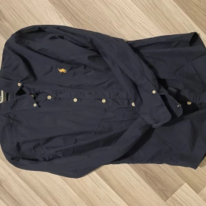 Mörkblå skjorta Polo Ralph Lauren S - Snygg mörkblå skjorta från Polo Ralph Lauren i slim fit. Klassisk design med knappar framtill och lång ärm, samt den ikoniska broderade loggan i gult på bröstet. Skjortan är gjord i mjuk bomull och passar perfekt till jeans eller chinos. Inga defekter!!