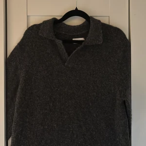 Grå stickad tröja från These Glory Days - Snygg grå stickad tröja från These Glory Days med krage och v-ringad öppning. Perfekt för kyliga dagar och har en klassisk, avslappnad look. Materialet är alpacka och känns mjukt och värmande, och tröjan har långa ärmar.