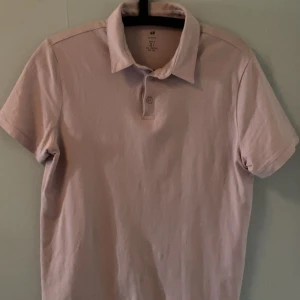 Ljusrosa pikétröja från H&M - Säljer en ljusrosa pikétröja från H&M i slim fit-modell. Tröjan har klassisk krage, korta ärmar och knäppning med två knappar framtill. Perfekt för en clean och stilren look.