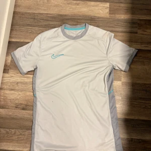 Vit Nike Dri-Fit fotbollströja M - Snygg vit fotbollströja från Nike med ljusgrå detaljer och turkos Nike-logga. Tröjan är i Dri-Fit-material som andas och har meshpaneler på sidorna för extra ventilation. Slim fit-modell med korta ärmar och rund hals.