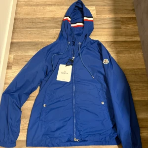 Blå vindjacka från Moncler med huva - Blå vindjacka från Moncler med huva och vita samt röda ränder på huvan. Jackan har dragkedja framtill, snörning i huvan och två fickor med tryckknappar. Tillverkad i lätt polyester, perfekt för blåsiga dagar. Klassisk Moncler-logga på ärmen.