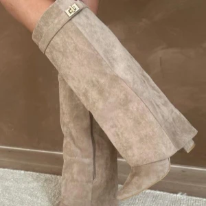 Beige mockastövlar med spetsig tå - Snygga höga stövlar i beige mocka med spetsig tå och låg klack. Stövlarna har en dekorativ rem med guldfärgat spänne upptill och dragkedja på insidan. Perfekta för att lyfta din outfit med en trendig touch.