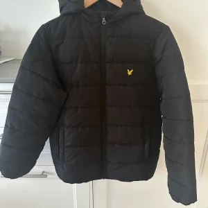 Svart pufferjacka från Lyle & Scott - Svart pufferjacka från Lyle & Scott med huva och gul logotyp på bröstet. Jackan har dragkedja framtill, två sidofickor och quiltad design. Perfekt för kalla dagar och har en stilren look som funkar till det mesta.