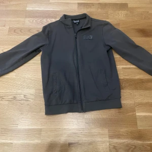 Mörkgrön ziptröja från EA7 Armani - Snygg mörkgrön ziptröja från EA7 Emporio Armani med diskret logga på bröstet. Tröjan har hel dragkedja, ståkrage och två fickor framtill. Perfekt för dig som gillar stilren och sportig look.