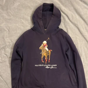 Marin blå hoodie Polo Bear Ralph Lauren S - Marin blå hoodie från Polo Ralph Lauren i storlek XL men det är barn storlek så i vuxen storlek är det ungefär XS/S med ikoniskt Polo Bear-motiv på bröstet där björnen rider häst och spelar polo. Tröjan har huva och är långärmad, tillverkad i mjuk bomull. Perfekt för dig som gillar streetstyle och klassiska märken.