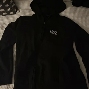 Svart EA7 Emporio Armani hoodie - Svart hoodie från EA7 Emporio Armani med dragkedja framtill och vit logga på bröstet. Tröjan har huva och är långärmad, perfekt för en chill och stilren look. Materialet är mjukt och bekvämt, troligen bomullsmix. Passar dig som gillar klassisk streetwear.