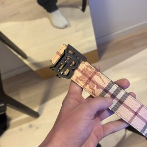 Burberry bälte - Säljer ett Burberry bälte i skinn med ikoniskt rutmönster i beige, brunt och rosa toner samt en stor svart metallspänne med Burberrys logotyp. Baksidan är helsvart med diskret rutstruktur. Perfekt accessoar för att lyfta din outfit.