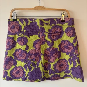 Blommig kortkjol  - Säljer en färgstark kortkjol med blommigt mönster i lila och rosa på en limegrön bakgrund. Kjolen har en rak passform och är tillverkad i ett mjukt, stickat material som känns bekvämt. Perfekt för dig som gillar att sticka ut med coola färger och mönster.