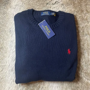 Mörkblå stickad tröja Polo Ralph Lauren - Klassisk mörkblå stickad tröja från Polo Ralph Lauren med rund halsringning och röd broderad logga på bröstet. Tröjan har ribbade muddar och är perfekt för en stilren look. Tillverkad i mjuk bomull för skön komfort.