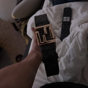 Svart Fendi bälte med guldspänne - Säljer ett svart bälte från Fendi med bred design och ikoniskt FF-mönster. Bältet har ett stort, fyrkantigt spänne i guldmetall som verkligen sticker ut. Tillverkat i skinn och perfekt för att lyfta vilken outfit som helst.