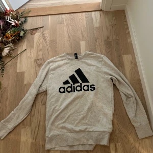 Beige sweatshirt från Adidas - Säljer en beige sweatshirt från Adidas med klassisk svart logga framtill. Tröjan har rund halsringning och långa ärmar. Perfekt för en sportig och avslappnad stil. Materialet är mjukt och skönt, troligtvis bomullsmix.