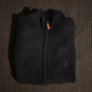 Mörkblå kofta från Ralph Lauren - Mörkblå kofta från Ralph Lauren med hel dragkedja och diskret broderad logga på bröstet. Tröjan har ribbade muddar och hög krage, perfekt för lager-på-lager. Klassisk och stilren design som funkar till det mesta.