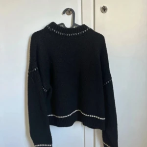 Svart stickad tröja med vita detaljer - Svart stickad tröja med vita sömmar vid hals, ärmar och nederkant. Tröjan har en avslappnad passform och långärmade ärmar, perfekt för kyliga dagar. Enkel och stilren design som funkar till många olika looks.