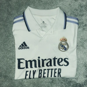 Real Madrid vit T-shirt  - Säljer en vit Real Madrid fotbollströja från Adidas med klubbmärke på bröstet. Tröjan har klassisk krage, korta ärmar och tre lila ränder på axlarna. Tillverkad i lätt och ventilerande material, perfekt för träning eller vardaglig användning. Kan gå ner i pris❗️