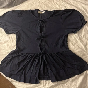 Mörkblå blus med knyt från Gina Tricot - Supersöt mörkblå blus från Gina Tricot med puffiga korta ärmar och tre knyt framtill. Blusen har rund hals och volang nedtill som ger en flowy känsla. Perfekt för dig som gillar feminina detaljer och vill ha en unik topp i garderoben. Pris kan diskuteras!😌
