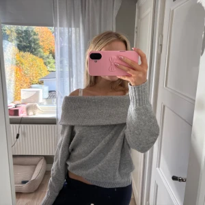 Grå offshoulder stickad tröja - Supermysig grå stickad tröja med bred offshoulder-krage och långa ärmar. Tröjan har en loose fit och ribbade muddar vid ärmslut och nederkant. Perfekt för kyliga dagar och enkel att styla med jeans eller kjol.