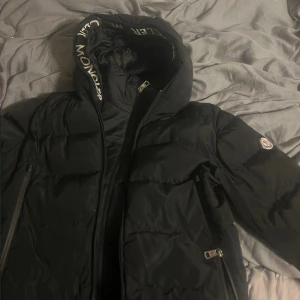 Svart pufferjacka från Moncler S - Säljer en svart pufferjacka från Moncler med huva och dragkedja. Jackan har logga på ärmen, stora vadderade partier och innerficka med röd dragkedja. Snyggt Moncler-tryck i luvan och klassisk design som håller dig varm under kalla dagar.