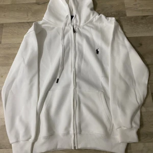 Vit hoodie från Polo Ralph Lauren - Vit hoodie från Polo Ralph Lauren med dragkedja framtill, snörning i huvan och klassisk broderad logga på bröstet. Mjuk bomullskvalitet och ribbade muddar. Perfekt för en clean och stilren look.