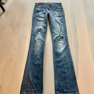 Blå bootcut jeans med slitningar  - Snygga blå bootcut jeans från Cross Jeans med slitningar på knäna och klassisk femficksdesign. Jeansen har låg midja, blå tvätt och coola detaljer på bakfickorna. Perfekta för dig som gillar en avslappnad och trendig look. Midjemåttet är ca 38 cm och innerbenslängden 84 cm