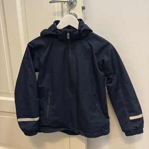 Blå Skaljacka från Kaxs Proxtec str 140 - Vind och vattentät blå skaljacka från Kaxs Proxtec str 140, med 10000mm vattenpelare. Jackan har huva, dragkedja framtill, två fickor med dragkedja och reflexdetaljer på ärmarna samt ett stort K på ryggen. Yttermaterial i polyamid och foder i polyester. Perfekt för blöta dagar. Har varit så nöjd med alla plagg vi köpt på Kappahl från Kaxs! I mycket fint skick! 