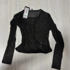 Svart glittrig mesh-top från New Yorker - Svart långärmad mesh-top med glittriga silverprickar över hela tyget. Tunn och genomskinlig, perfekt för att sticka ut. Rund halsringning och figurnära passform. Perfekt inför nyår! 