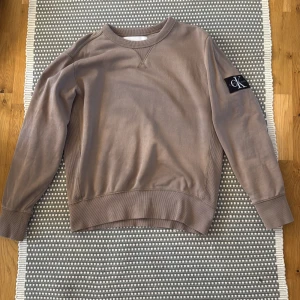 Beige sweatshirt från Calvin Klein Jeans - Säljer en beige sweatshirt från Calvin Klein Jeans med klassisk rund hals och ribbade muddar. Tröjan har en svart CK-logga på ena ärmen och är i mjuk bomull. Perfekt för en chill och stilren look.