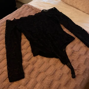 Svart spetsig offshoulder bodysuit - Säljer en svart bodysuit med långärmade spetsärmar och offshoulder-design. Toppen har en tight passform och är tillverkad i genombruten spets som ger en snygg och edgy look. Perfekt för dig som vill sticka ut med coola detaljer och feminin stil.