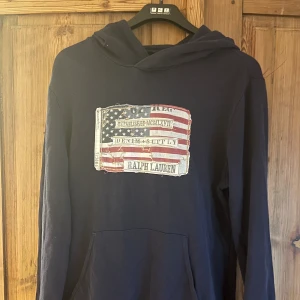 Mörkblå hoodie från Ralph Lauren - Snygg mörkblå hoodie från Ralph Lauren med stor amerikansk flagga och broderad text på bröstet. Tröjan har huva, 